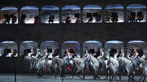 Eine Gruppe von Reiterinnen auf Schimmeln, begleitet von einem Orchester in einer Arena.