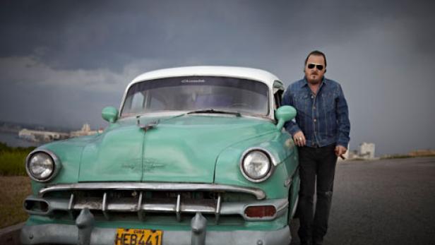 Ein Mann lehnt an einem türkisfarbenen Oldtimer vor einer Skyline unter einem bewölkten Himmel.