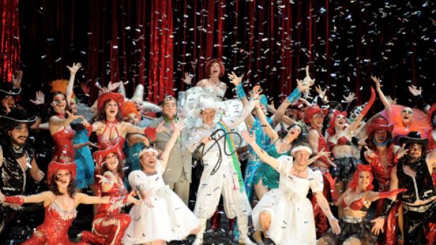 Eine Gruppe von Darstellern in bunten Kostümen feiert auf einer Bühne unter fallendem Konfetti.