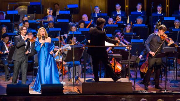 Ein Orchester spielt mit einem Sänger, einer Sängerin und einem Violinisten auf einer Bühne.