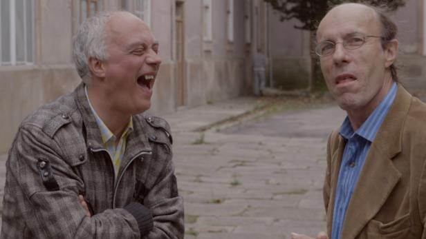 Ein Mann lacht, während ein anderer mit Brille ihn ansieht.