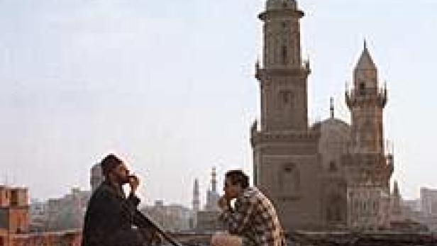 Zwei Männer sitzen auf einem Dach mit Blick auf eine Moschee in einer Stadt.