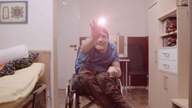 Ein junger Mann im Rollstuhl winkt in einem hellen Raum.