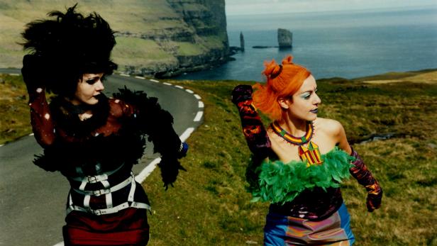 Zwei Frauen in auffälligen Kostümen posieren auf einer Straße vor einer Küstenlandschaft.