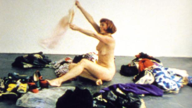 Eine Frau sitzt inmitten eines Haufens Kleidung und hält ein Kleidungsstück hoch.