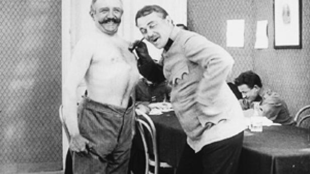 Ein Mann mit freiem Oberkörper und ein uniformierter Mann stehen nebeneinander.