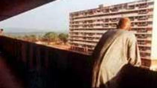 Ein Mann steht auf einem Balkon und blickt auf ein Hochhaus und die Landschaft.