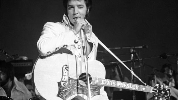 Elvis Presley tritt in einem weißen Anzug mit Gitarre auf einer Bühne auf.