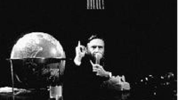 Ein Mann mit Bart sitzt an einem Tisch mit einem Globus und hält ein Glas in der Hand.