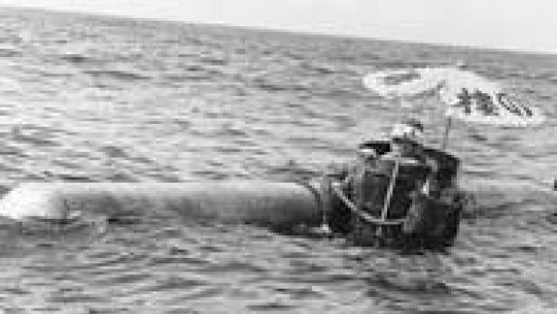 Ein Mann treibt mit einem ungewöhnlichen, rohrartigen Floß und einem Sonnenschirm auf dem Meer.