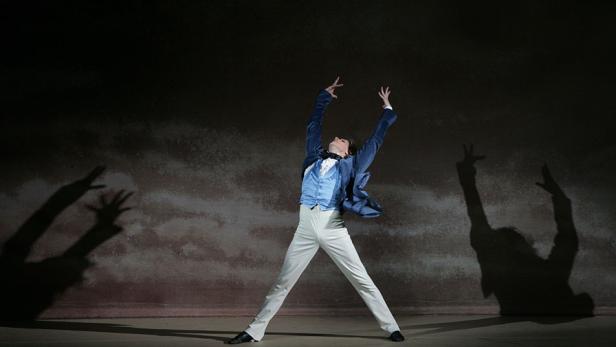 Ein Tänzer in einem blauen Anzug steht mit erhobenen Armen auf einer Bühne.