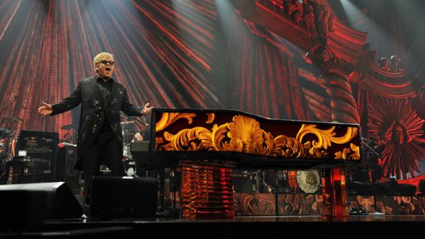 Elton John steht auf einer Bühne mit einem verzierten Klavier und singt.