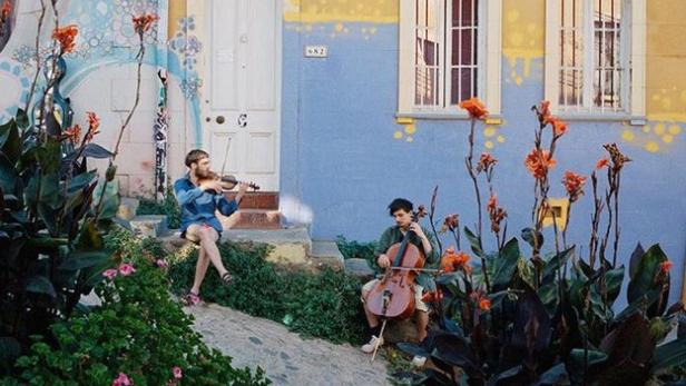 Zwei Musiker spielen Geige und Cello vor einem farbenfrohen Haus.