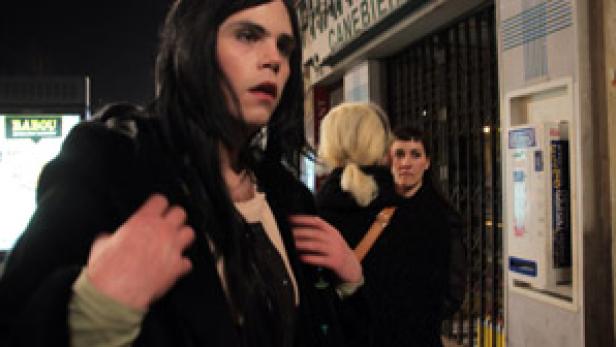 Ein junger Mann mit dunklen, langen Haaren und dunkler Jacke steht nachts auf der Straße.