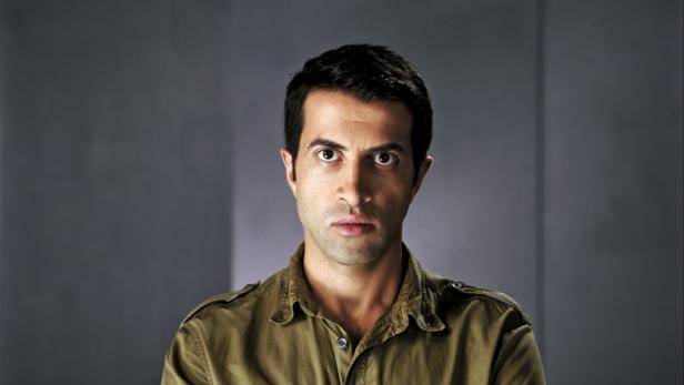 Porträt eines Mannes mit dunklen Haaren und einem olivgrünen Hemd vor einem grauen Hintergrund.