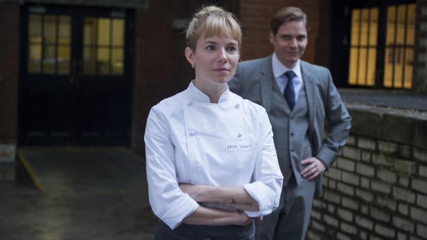 Sienna Miller als Köchin mit verschränkten Armen, Daniel Brühl im Hintergrund.