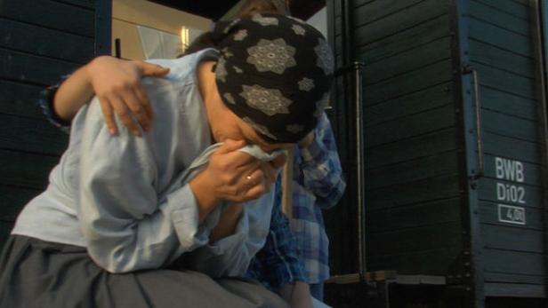 Eine Frau weint, während sie von einer anderen Person getröstet wird; im Hintergrund ein Güterwagen.