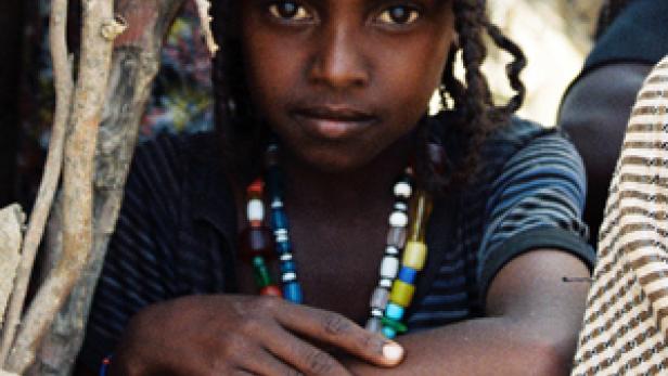 Ein junges Mädchen mit geflochtenen Zöpfen und einer bunten Halskette sitzt im Freien.