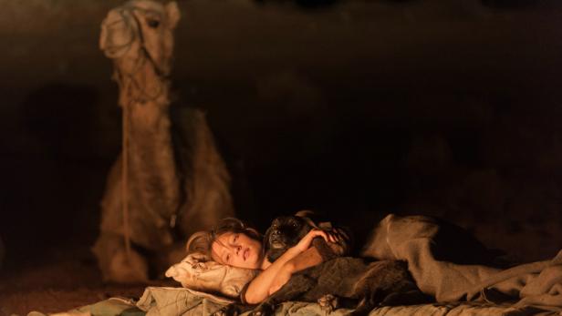 Eine Frau liegt mit einem Hund in der Wüste, im Hintergrund ein Kamel.