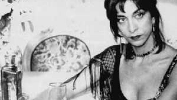 Schwarzweißporträt von Lydia Lunch mit einem Glas und einer Flasche auf dem Tisch.
