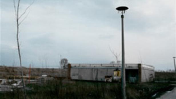 Ein kleines, graues Gebäude steht unter einem bewölkten Himmel neben einer Straßenlaterne.