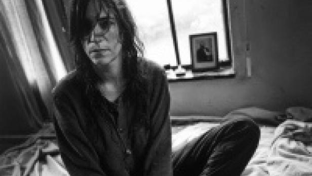 Schwarzweißporträt von Patti Smith, die auf einem Bett vor einem Fenster sitzt.