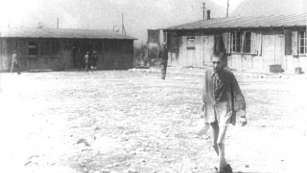 Ein Mann in Shorts und Hemd geht über einen Platz mit Baracken im Hintergrund.