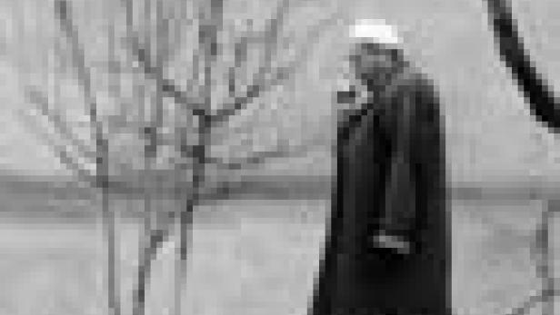 Ein älterer Mann mit Mantel und Mütze steht draußen und raucht.