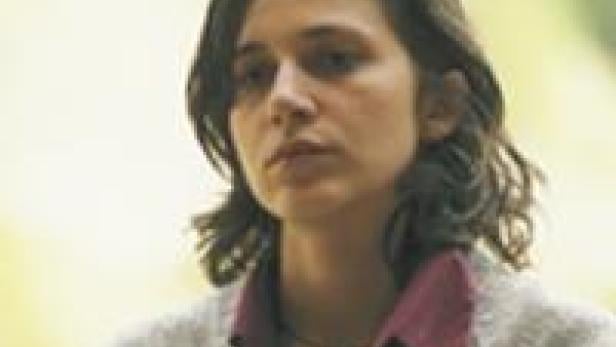 Eine Frau mit dunklen, lockigen Haaren und einem ernsten Gesichtsausdruck.