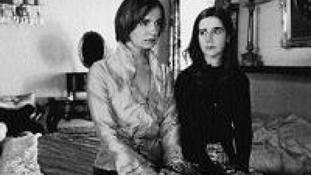 Zwei Frauen sitzen in einem Schlafzimmer, die eine trägt eine Bluse, die andere einen Pullover.