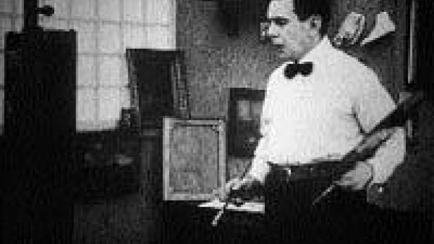 Ein Mann mit Fliege steht in einem Atelier und hält Pinsel und Palette.