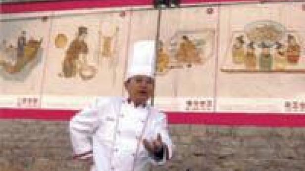 Ein Koch in Kochuniform steht vor einer Wand mit historischen Malereien.