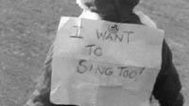 Ein Kind trägt ein Schild mit der Aufschrift „I WANT TO SING TOO!“.