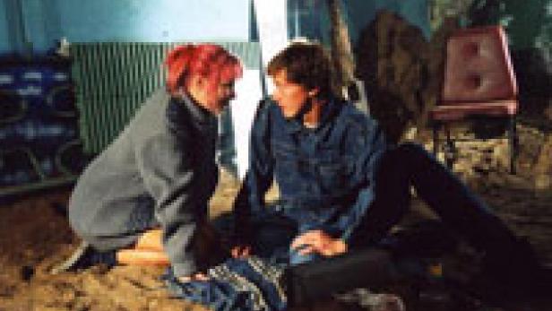 Eine junge Frau mit pinken Haaren und ein junger Mann sitzen auf dem Boden und schauen sich an.