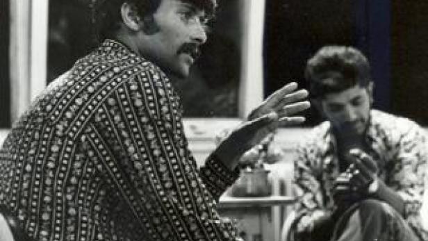 Ein Mann mit Schnurrbart gestikuliert, während ein anderer Mann im Hintergrund sitzt.