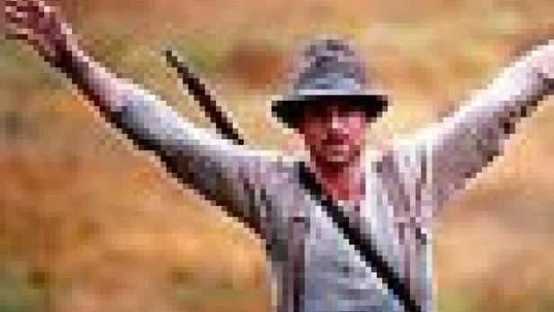 Ein Mann mit Hut und Gewehr steht mit erhobenen Armen in der Natur.