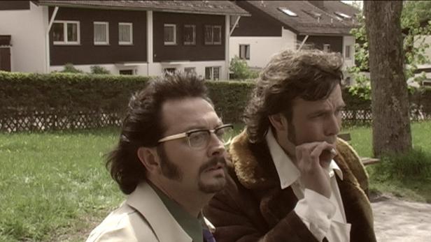 Zwei Männer mit Schnurrbärten und Retro-Kleidung stehen im Freien.