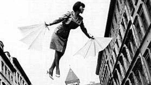 Eine Frau „fliegt“ mit Hilfe von zwei geöffneten Regenschirmen zwischen Häusern hindurch.