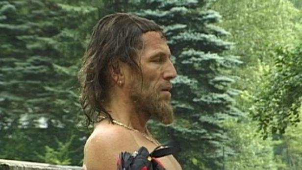 Ein Mann mit Bart und nacktem Oberkörper sitzt auf einer Bank vor Bäumen.