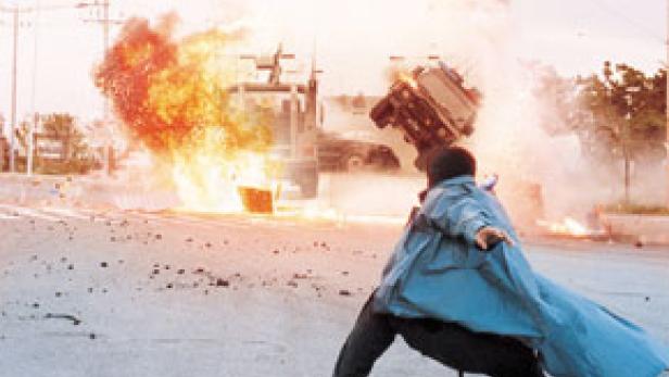 Ein Mann wirft einen Gegenstand auf ein Auto, das explodiert.