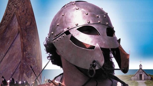 Ein Mann mit Wikingerhelm steht vor einem Wikingerschiff und einer kleinen Kirche am Meer.