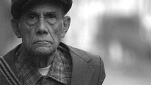 Ein älterer Mann mit einer Schiebermütze und kariertem Hemd blickt ernst in die Kamera.