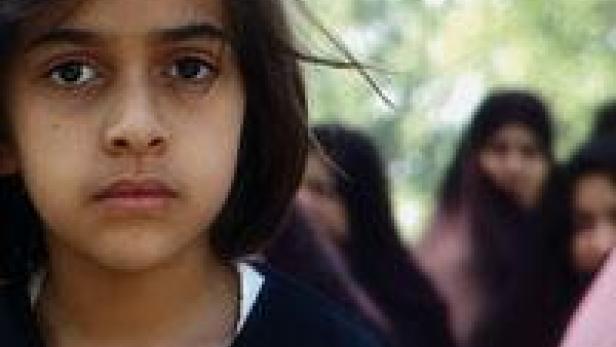 Ein ernstes junges Mädchen steht vor einer Gruppe von Frauen mit Kopftüchern.