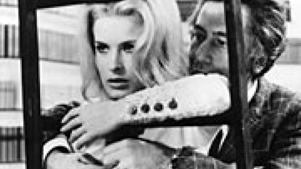 Eine blonde Frau wird von einem Mann umarmt, beide stehen hinter einem dunklen Rahmen.