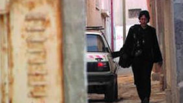 Eine Frau geht eine enge Gasse entlang, im Hintergrund ein geparktes Auto.