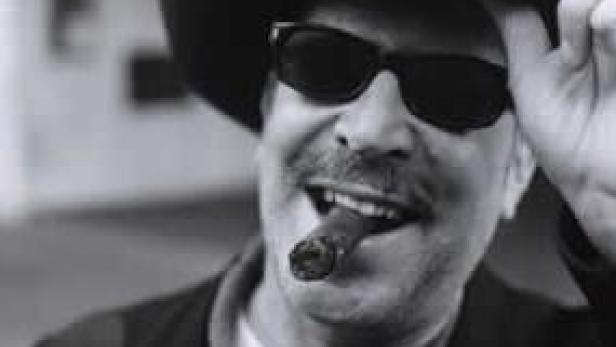 Ein Mann mit Hut, Sonnenbrille und Zigarre lächelt.