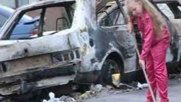 Ein Mädchen kehrt Trümmer vor einem ausgebrannten Auto.