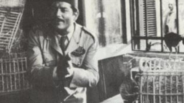 Ein Mann mit Hut und Uniform hält einen Vogel in der Hand, umgeben von Vogelkäfigen.