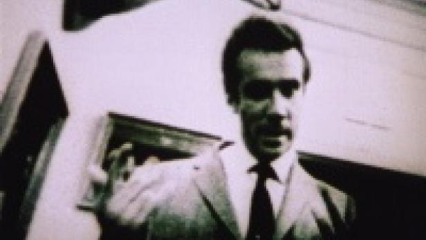 Ein Mann in Anzug und Krawatte gestikuliert vor einer Wand.