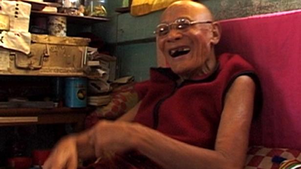 Ein älterer, glatzköpfiger Mann mit Brille lacht, während er auf einem Bett sitzt.